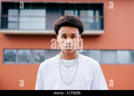 Young black curly haired male teenager in stylish clothes with chains looking at camera while standing on city street with buildings in background Stock Photo