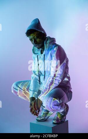 Handsome African American male in trendy outfit looking at camera while sitting on haunches on cube under colorful light Stock Photo