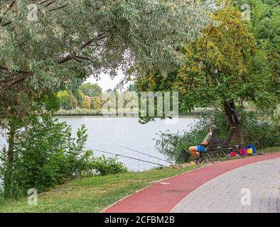 Fisherman fishing at lake sitting on chair under the cherry plum tree. Two fishing rods mounted on the holder and set up at the shore of a lake. Beaut Stock Photo