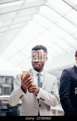 afroamerican bearded Businessman wearing suit and glasses texting on phone in mall or hall of modern office. Business Concept Stock Photo