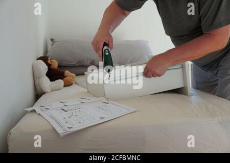 A man assembling a drawer of a computer desk for his son using power cordless screwdriver. He is using a tool and instructions to complete the project. Stock Photo