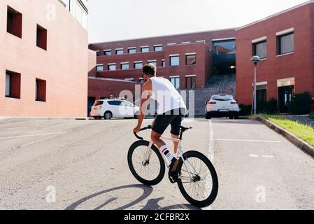 Sportive male in sunglasses wearing white sleeveless shirt and black shorts standing while riding a bicycle between buildings connected on summer sunny day Stock Photo