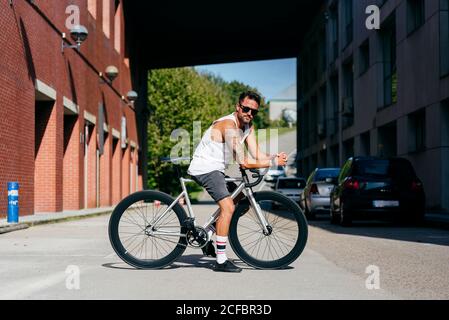 Sportive male in sunglasses wearing white sleeveless shirt and black shorts sitting on bicycle between buildings connected by bridge on summer sunny day Stock Photo