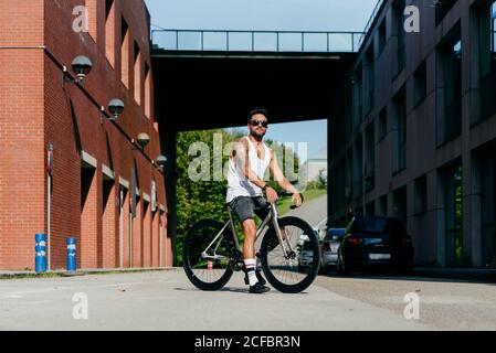 Sportive male in sunglasses wearing white sleeveless shirt and black shorts sitting on bicycle between buildings connected by bridge on summer sunny day Stock Photo