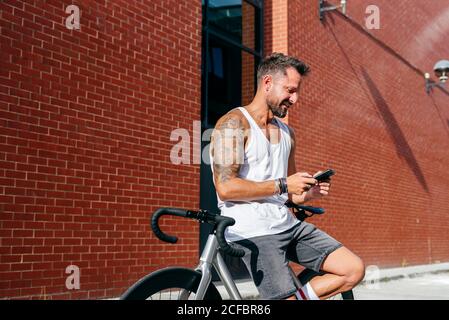 Handsome male cyclist in sportswear using smartphone while resting on bike next to red brick wall Stock Photo