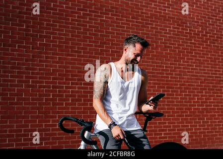 Handsome male cyclist in sportswear using smartphone while resting on bike next to red brick wall Stock Photo