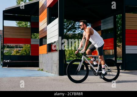 Sportive male in sunglasses wearing white sleeveless shirt and black shorts standing while riding a bicycle between buildings connected on summer sunny day Stock Photo