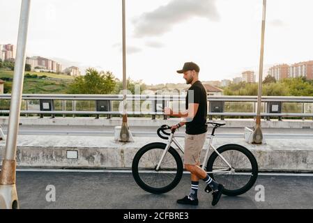 Side view of young casual bearded male cyclist in black cap using phone while walking with bike on road with city buildings on background Stock Photo