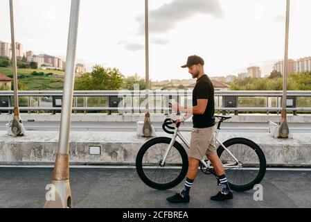 Side view of young casual bearded male cyclist in black cap using phone while walking with bike on road with city buildings on background Stock Photo