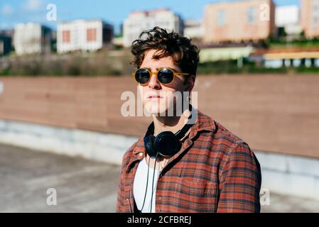 Handsome young man in checkered shirt and sunglasses looking at camera while standing on blurred background of city street Stock Photo