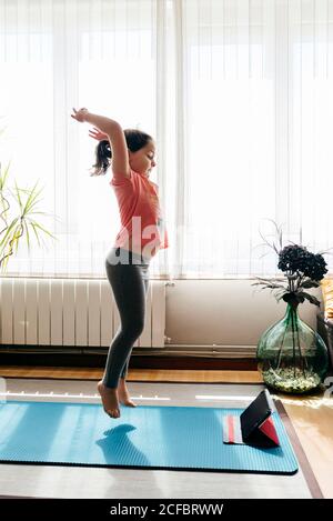 Side view of full length female kid in casual wear jumping on mat against window at home while doing yoga and watching video tutorials on tablet Stock Photo