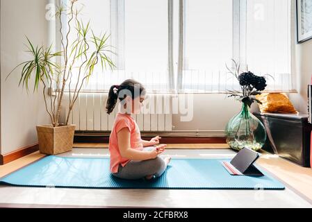 Little girl sitting in lotus pose and meditating on mat with eyes closed at home with big window while practicing yoga and watching video tutorials on tablet Stock Photo