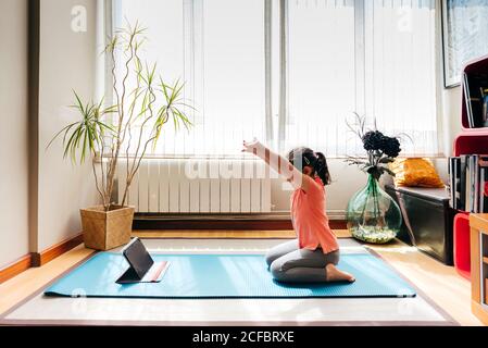 Side view of positive little girl sitting on her knees in yoga pose on mat near window at home while practicing yoga and watching video tutorials on tablet Stock Photo