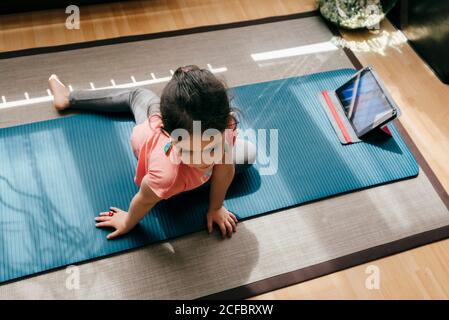 From above positive little girl in yoga pose on mat near window at home while practicing yoga and watching video tutorials on tablet Stock Photo