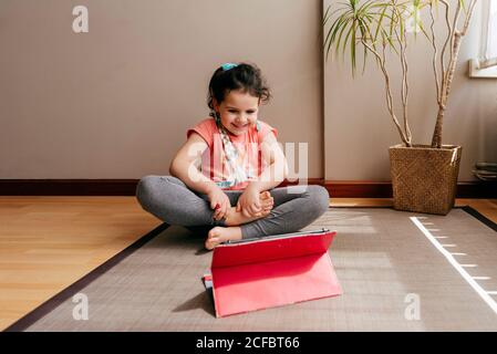 Full body little girl sitting in lotus pose and meditating on mat at home with big window while practicing yoga and watching video tutorials on tablet Stock Photo