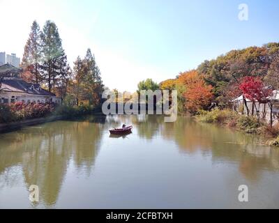 A lonesome boat with a dummy sitting in it floats on a river located inside the Korean Folk Village surrounded by thick fall foliage of various primar Stock Photo