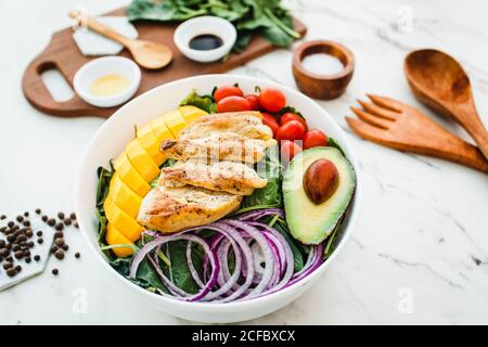 From above fried chicken and sliced mango placed near cherry tomatoes and half of avocado on spinach leaves and onion on marble table near utensils and sauces with spices Stock Photo