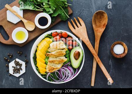 Top view of fried chicken and sliced mango placed near cherry tomatoes and half of avocado on spinach leaves and onion on gray marble table near utensils and sauces with spices Stock Photo