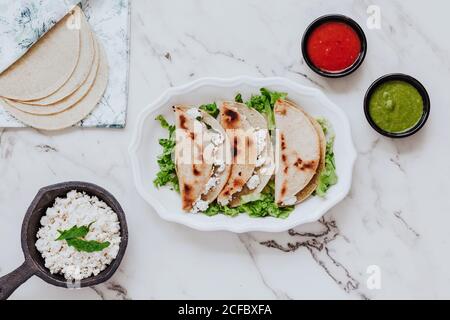Top view of tortillas with fresh quark cheese placed on plate with lettuce leaves near bowls of avocado and tomato sauces on marble table Stock Photo