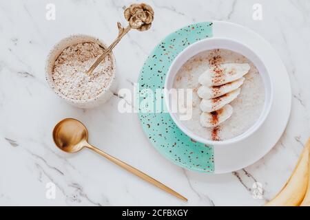 Top view of bowl of healthy oatmeal porridge served with slices of banana and cinnamon near spoon on marble tabletop Stock Photo