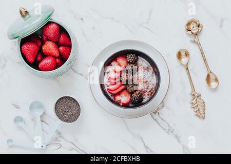 From above slices of ripe strawberries and blackberries in cinnamon powder in yogurt bowl in composition with bowl with fresh whole berries and golden teaspoons on white table Stock Photo