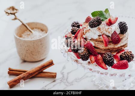 Tasty pancakes with fresh strawberries and blackberries decorated with mint leaf placed on plate near cinnamon sticks and cup of ice cream on marble table Stock Photo