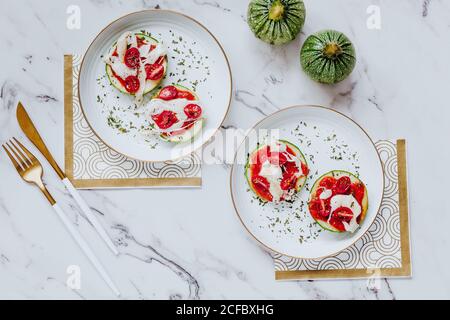 From above delicious healthy sandwiches with slices of fresh green zucchini with melted mozzarella and cherry tomatoes on white plates with golden cutlery Stock Photo