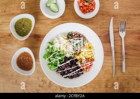 Top view of lettuce and beans with chopped tomatoes placed near corn kernels and grain in bowl on wooden table near various sauces with lime and silverware Stock Photo