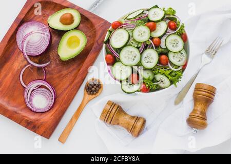 Flat lay of bowl with salad of cucumber and lettuce with red onion and cherry tomatoes on white table with board and seasonings around Stock Photo
