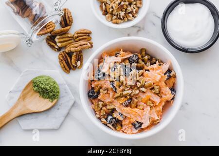 Top view of big white bowl with slices of carrot in yogurt dressing with pecan nuts and prunes on marble table Stock Photo