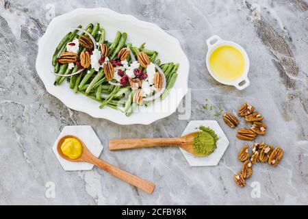 From above of oval plate with salad of green beans topped with goat cheese and pecan nuts on marble surface with dressing ingredients Stock Photo