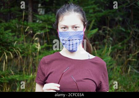 Portrait of young woman with brown hair ponytail in a colorful patterned face mask on a nature background Stock Photo