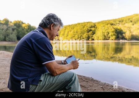 Side view of senior man using mobile phone while sitting amazing lake mountain landscape Stock Photo