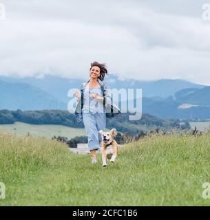 Smiling running beagle dog portrait with tongue out and happy owner female jogging by the mounting meadow grass path. Walking in nature with pets, hap Stock Photo