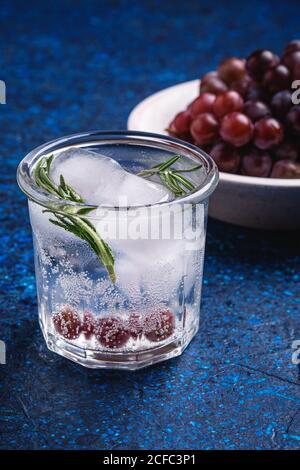 Fresh ice cold carbonated water in glass with rosemary leaf near to wooden bowl with grape berries, blue textured background, angle view macro