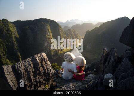 From above back view of couple of tourists in conical hats hugging while sitting on high rocky top of Mua cave and admiring magnificent tropical valley in ridges