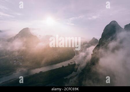 Drone view of calm river and rocky dark mountains in morning mist with flowing clouds under bright sky and sun