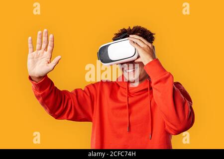 Freestyle. Mulatto guy in hoodie and vr headset standing isolated on yellow watching video touching space cheerful Stock Photo