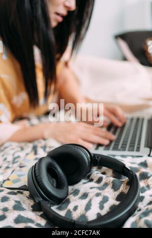 Side view of young female in home clothes sitting on comfortable bed with laptop and close a headphones while resting in cozy bedroom at home Stock Photo
