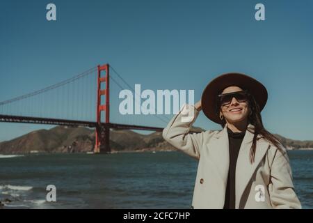 Side view of smiling young Woman in trendy outfit with hat and sunglasses standing on embankment against Golden Gate Bridge in California in sunny day Stock Photo
