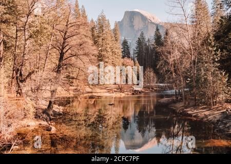 Spectacular peaceful landscape of calm river flowing through coniferous forest in mountain valley with rocky cliff reflected in sunny spring day in Yosemite National Park in California Stock Photo