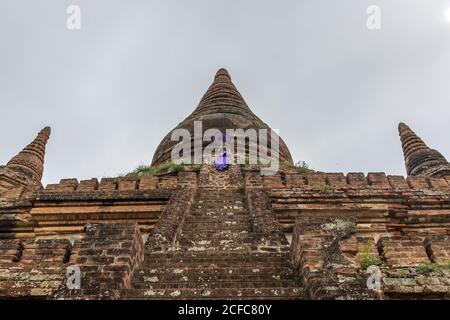 From below of ancient Burmese stone temple in Bagan against grey cloudy sky with female in purple dress holding purple umbrella standing on stairs Stock Photo