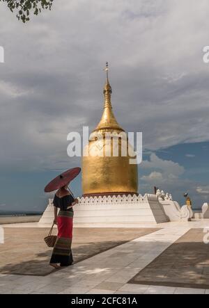 Back view of female in traditional long dress holding red umbrella and bag walking next to beautiful golden bell shaped Burmese stupa in Bagan Myanmar Stock Photo