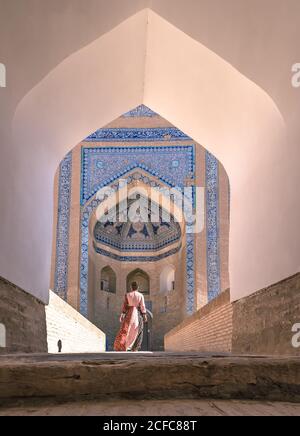 Back view of female in colorful dress walking along street against old building with arch and blue oriental mosaic on walls in Khiva in fair weather Stock Photo