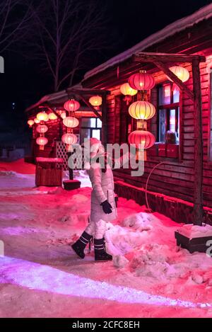 Side view of young female in outwear examining bright Chinese lanterns while standing outside village house at winter night Stock Photo