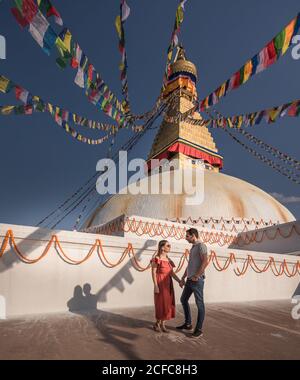 Couple holding hands and looking at each other while standing near Buddhist temple with decorative garlands and tower under cloudy sky in daylight Stock Photo