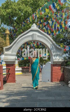 Female tourist in dress looking away while standing on pavement walking on old Buddhist temple under colorful garland with flags on sunny day Stock Photo
