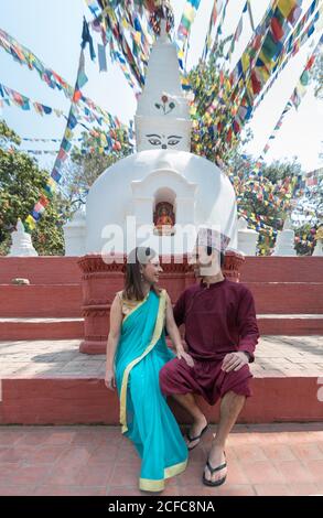Couple holding hands and looking at each other while sitting on red stairs near ancient sculpture and arch under garland with prayer flags Stock Photo