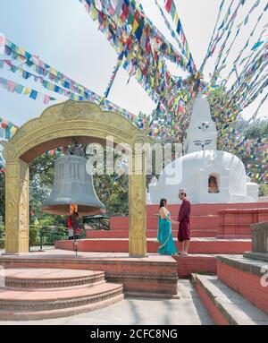 Side view couple holding hands and looking at each other while standing on red stairs near ancient sculpture and arch with big bell under garland with prayer flags Stock Photo