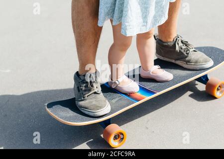 faceless man in sneakers and little girl in dress standing together on skateboard Stock Photo